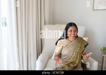 Senior indian asian woman wearing sari sitting at home on armchair with artwork Stock Photo