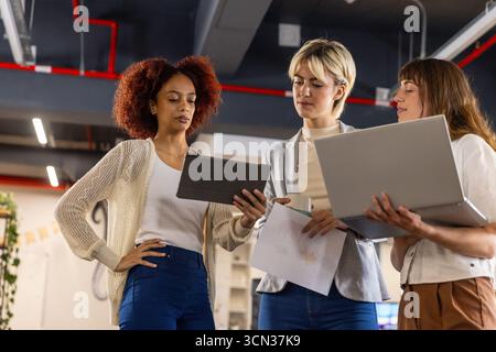 Diverse female colleagues standing and working on project in modern office with tablet and document Stock Photo