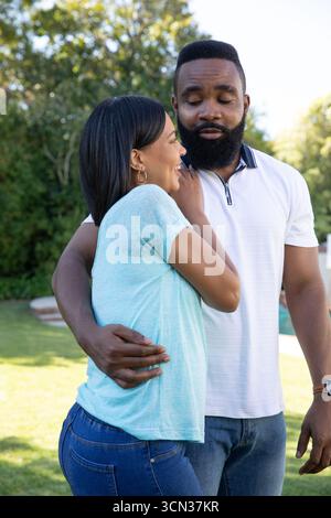 Diverse couple embracing beside swimming pool in sunlit backyard showing wristwatch hoop earrings Stock Photo