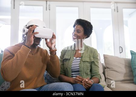African american couple sitting on couch at home, adjusting white vr headset while woman smiling Stock Photo