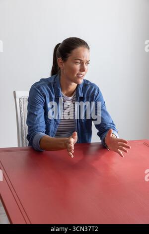 Woman gesturing with both hands explaining point at red tabletop inside room on chair Stock Photo
