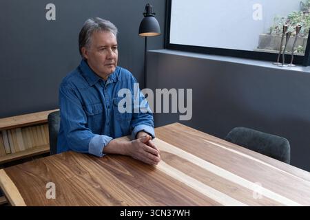 Senior man sitting at table with black lamp gazing at window in home, copy space Stock Photo