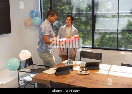 Diverse coworkers standing by table in office holding gift box amid balloons, cakes, copy space Stock Photo