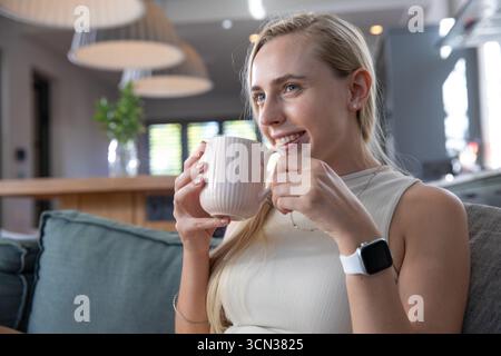 Woman holding ribbed ceramic mug next to face and gazing right in open-plan living room, copy space Stock Photo