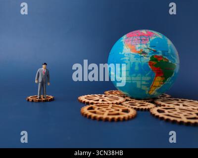The miniature businessman standing on a wooden gear, with toys globe on a blue background. Stock Photo