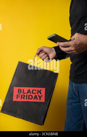 Customer using smartphone and holding shopping bag with black friday inscription against yellow background Stock Photo