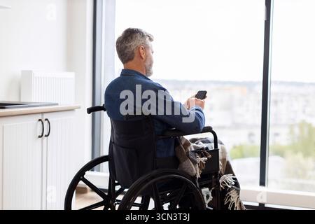 Man in wheelchair using smartphone indoors looking out large window view Stock Photo