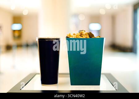 A dark blue disposable beverage cup with a lid stands next to a teal popcorn box filled with popcorn and dark pieces, placed on a white surface framed Stock Photo
