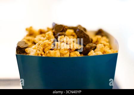 Close-up shot of popcorn kernels mixed with dark chocolate pieces, served in a blue square-shaped paper container. The background is blurred and white Stock Photo