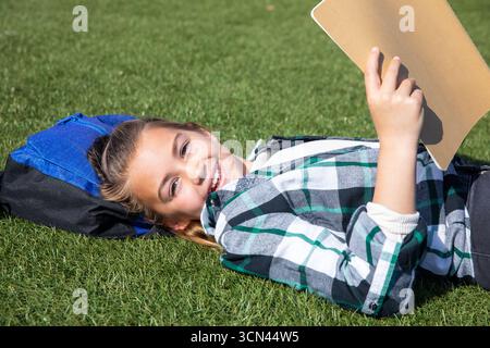 Female child lying on grassy field using blue and black backpack as pillow with tan clipboard Stock Photo