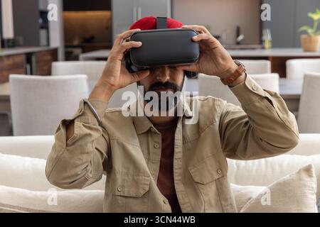 Bearded indian man wearing red turban lifting black vr headset on light-colored couch, kitchen Stock Photo
