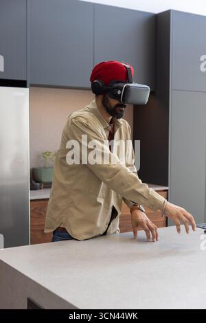 Man wearing red turban with vr headset extending hands over stone counter in kitchen by fridge Stock Photo