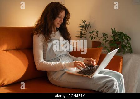 Pregnant woman sitting comfortably on an orange sofa, working remotely on a laptop in her cozy living room, balancing career and motherhood during her pregnancy journey Stock Photo