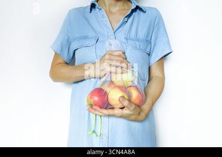 Woman standing against a blank background holds apples in a mesh bag. Eco-conscious shopper. Zero plastic, clean eating habits, local produce support Stock Photo