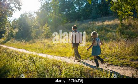 Boy with backpack and girl in blue dress walking on forest trail in sunlight Stock Photo