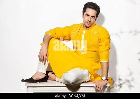 A stylish South Asian man in a vibrant yellow kurta and white churidar poses with a confident gaze while sitting on a stool in a studio. Stock Photo