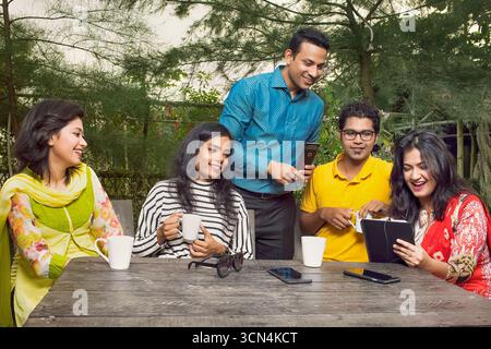 A diverse group of five friends, a mix of men and women, are gathered around a wooden outdoor table. They are enjoying coffee and conversation. Stock Photo