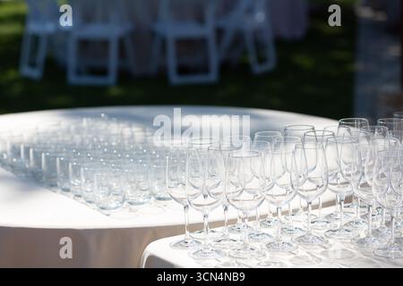 Rows of gleaming empty wine glasses and tumblers stand ready on tables with white tablecloths at an outdoor event, awaiting guests and refreshments Stock Photo