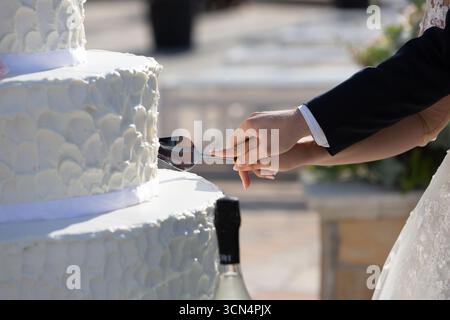 Newlyweds cutting their white, tiered wedding cake with a cake server at an outdoor reception Stock Photo