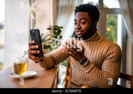 A young black man sits at the bar, casually dressed. He is using smartphone and having video call. Stock Photo