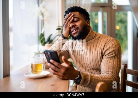 A young black man sits at the bar, using smartphone, messaging online. He is anxious and preoccupied. Stock Photo