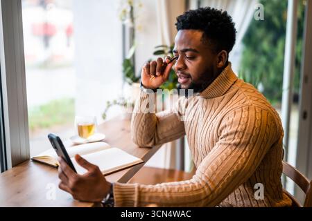Young black man sitting in bar. He is using smartphone and notepad. Freelancer making business plans. Stock Photo