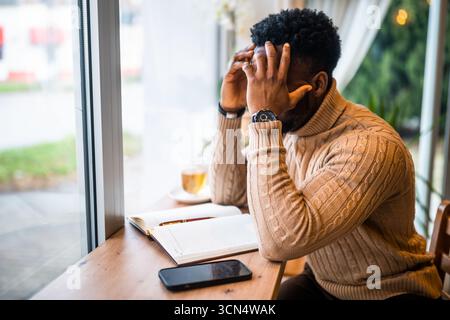 Young black man sitting in bar. He is using smartphone and notepad. Freelancer making business plans. Stock Photo