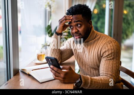 Young black man sitting in bar. He is using smartphone and notepad. Freelancer making business plans. Stock Photo