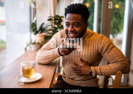 A young black man sits at the bar, casually dressed. He is using smartphone and recording voice message. Stock Photo