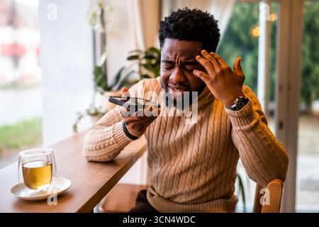 A young black man sits at the bar, casually dressed. He is using smartphone and recording voice message. Stock Photo
