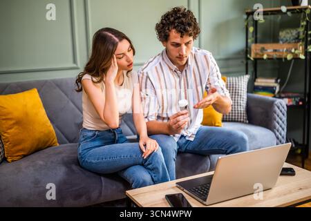 Adult couple sitting on bed in living room and having online doctor's appointment using laptop. Woman having headache. Remote consulting with doctor. Stock Photo