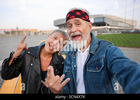 Stylish senior couple showing rock gesture while taking selfie near modern cabriolet outdoors Stock Photo