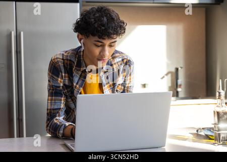 Male student sitting at kitchen working on silver laptop wearing plaid shirt and earbuds Stock Photo