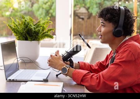 Man wearing red shirt and headphones speaking into microphone at table with laptop Stock Photo
