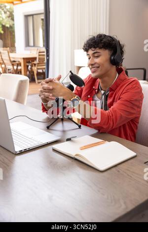 Hispanic man using usb microphone with headphones at home desk working on laptop Stock Photo