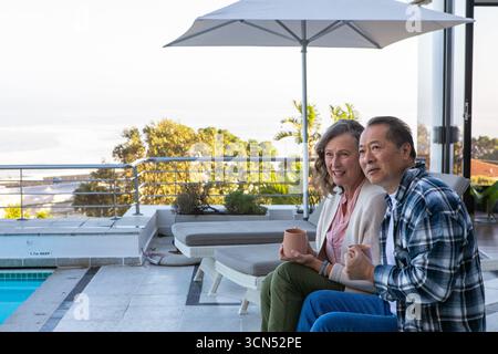 Diverse senior couple relaxing at poolside holding mugs near umbrella with ocean view, copy space Stock Photo