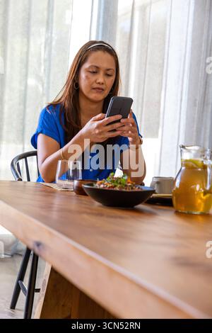 Asian woman sitting at dining table in cafe holding smartphone beside salad bowl, water glass Stock Photo