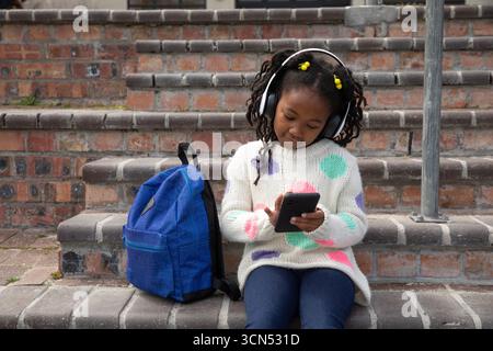 African american girl sitting on staircase holding tablet in headphones beside blue backpack Stock Photo