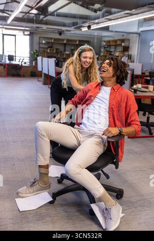 Diverse coworkers laughing while rolling office chair over scattered papers in open office space Stock Photo