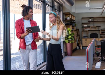 Diverse coworkers walking along glass walls at office, viewing tablet and pointing at screen Stock Photo