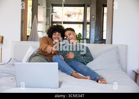 Diverse male partners using silver laptop while sitting on king-size bed in modern bedroom Stock Photo