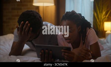 Displeased shocked African American family couple boyfriend man girlfriend woman lying bed use online digital tablet pad browsing internet lost Stock Photo