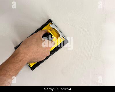 A builder smooths a wall after plastering using a sanding tool. The image depicts construction work and interior finishing in home improvement. Stock Photo