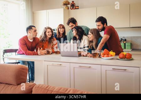 A diverse group of young friends relax and chat in a modern kitchen, using a laptop to plan something together while enjoying drinks and snacks at hom Stock Photo