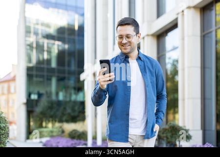 Young man in glasses and casual clothes smiling while walking downtown, holding smartphone and browsing messages and apps in front of a modern office building during daytime Stock Photo