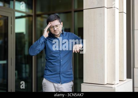 Young man standing outside building feeling painful headache or migraine, pressing hand against temple with eyes closed, experiencing stress and discomfort Stock Photo