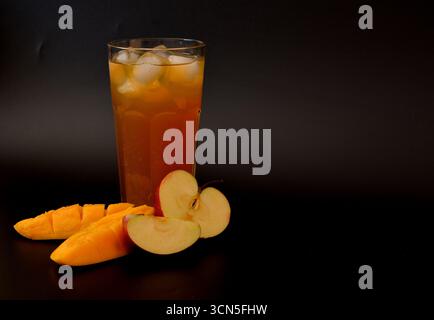 A mixture of red apple juice and tropical mango fruit juice in a tall faceted glass with ice cubes on a black background, next to ripe sliced ​​fruits Stock Photo