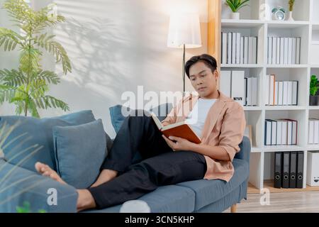 A young man sits comfortably on a sofa reading a book, appearing thoughtful and focused. The cozy indoor setting highlights relaxation, concentration, Stock Photo