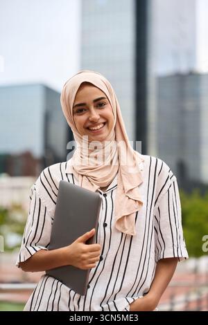 Young muslim woman smiling holding laptop in city Stock Photo