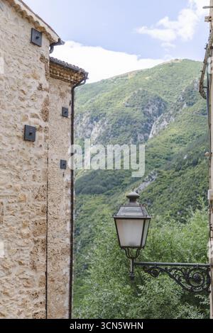 View of the old stone buildings framing a lush green mountain range under a bright sky, a vintage street lamp casting shadows, Castrovalva, Abruzzo, Italy. Stock Photo